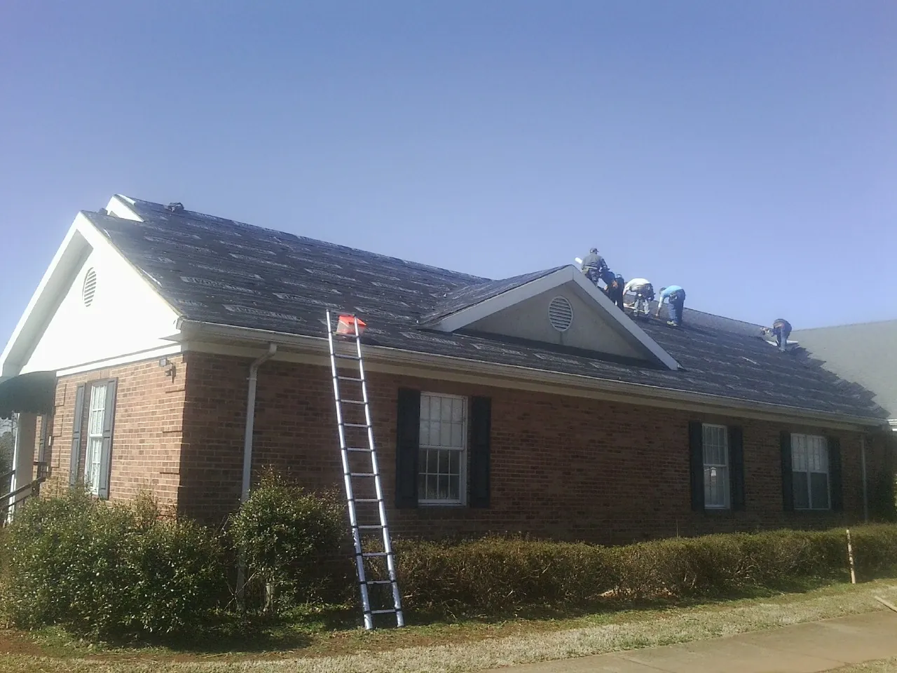 Crew tearing shingles off a brick ranch roof with a ladder set — Metal Roof Repair work by our crew in Eldena